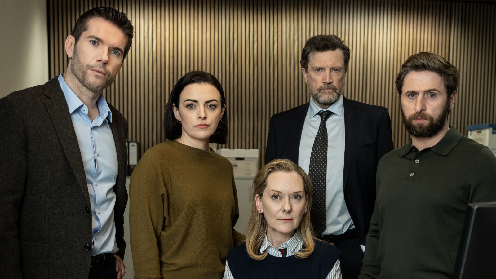 A man, woman, seated woman, and two other men stare straight out of the frame in an office setting.